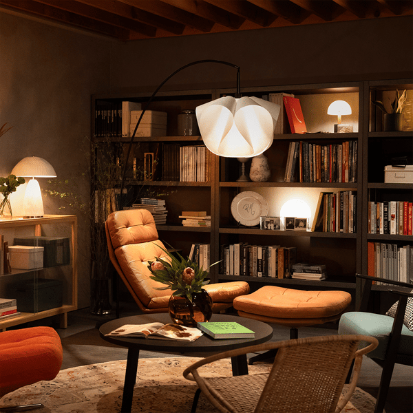 本棚と間接照明に囲まれたリビングの一角。キャメル色のラウンジチェアとオットマン、丸テーブルに花瓶と本が置かれたくつろぎスペース。
