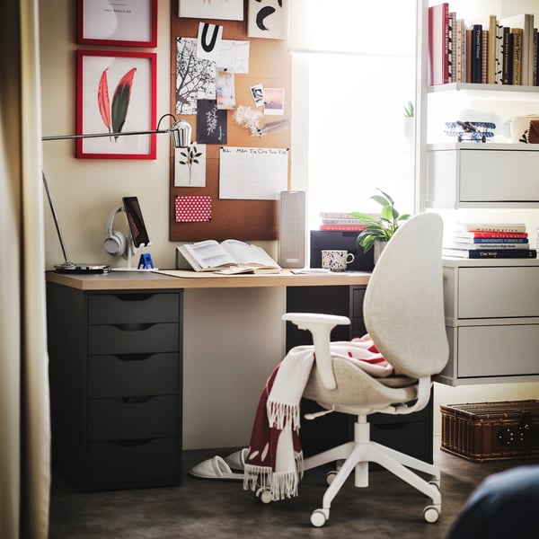 A white SYMFONISK WiFi bookshelf speaker on top of a cosy desk in a bright room with a beige HATTEFJÄLL armchair in front.