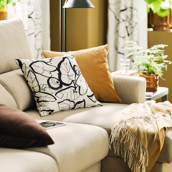 Cushions and a throw cover a light-beige MALMKÖPING sofa with chaise-longue, next to a TINGBY side table holding a plant.
