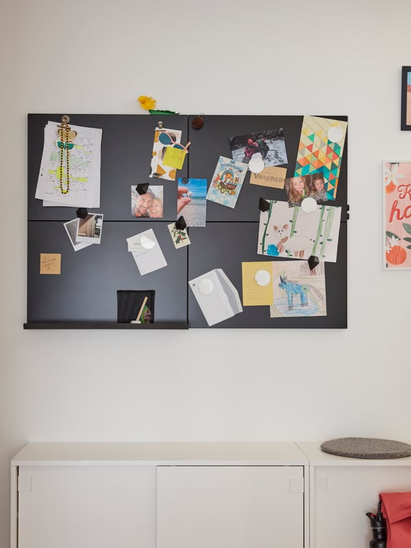 A black memo board with drawings, photos and magnets. A white bench with storage is below.