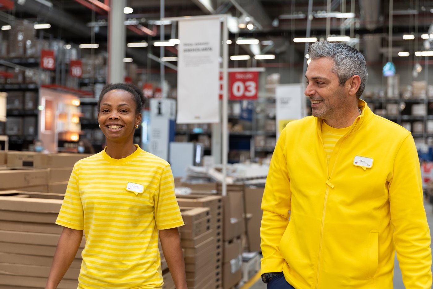 Kaksi IKEA-työntekijää kävelee yhdessä IKEA-tavaratalon itsepalveluvaraston halki hymyillen keltaisissa IKEA-työasuissaan.