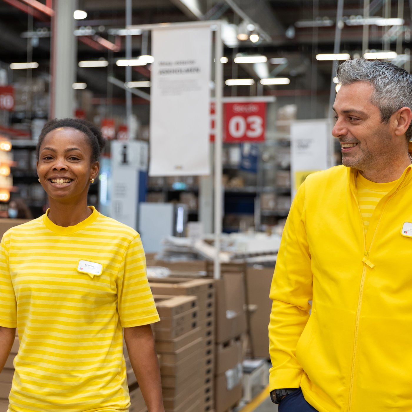 Kaksi IKEA-työntekijää kävelee yhdessä IKEA-tavaratalon itsepalveluvaraston halki hymyillen keltaisissa IKEA-työasuissaan.
