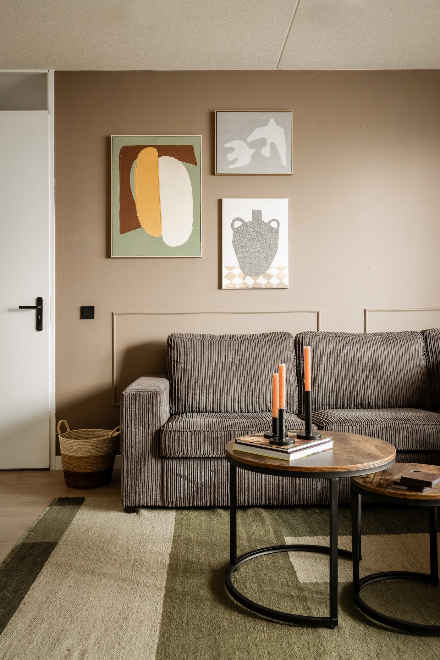 Woonkamer met een lichtbruine tint op de muur, waaraan ook drie schilderijen hangen. Er staat een bruine ribstof bank op een groen-beige gestreept vloerkleed, voor de bank staan twee ronde salontafels met daarop kaarsen.