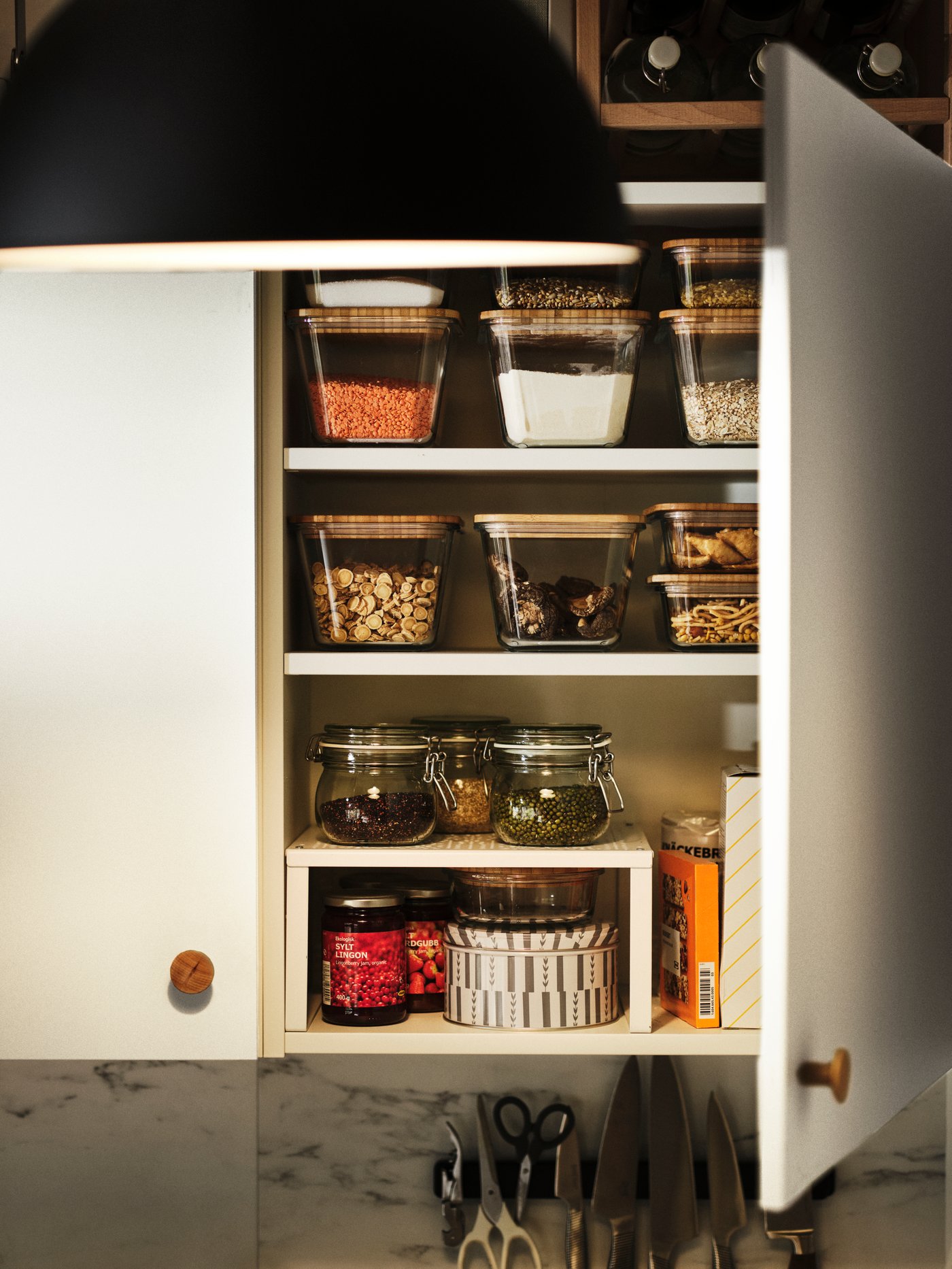 Estante adicional VARIERA con tarros y tapas encima, colocado en un armario con dos estantes junto a recipientes de alimentos.