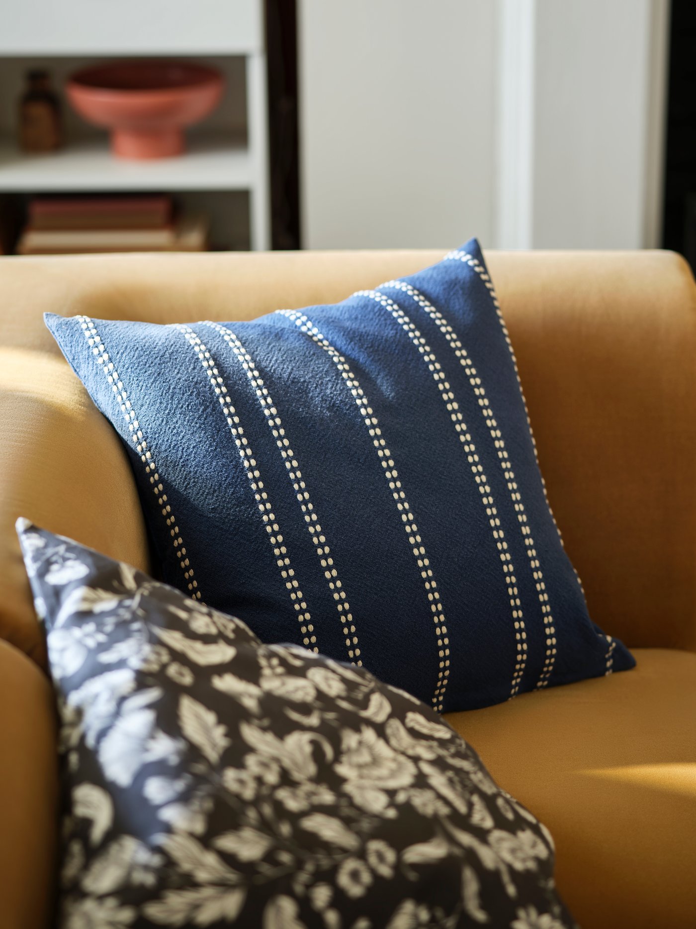 A striped KRUTBRÄNNARE cushion cover in blue off-white sits on a beige STOCKHOLM two-seat sofa next to a patterned cushion.