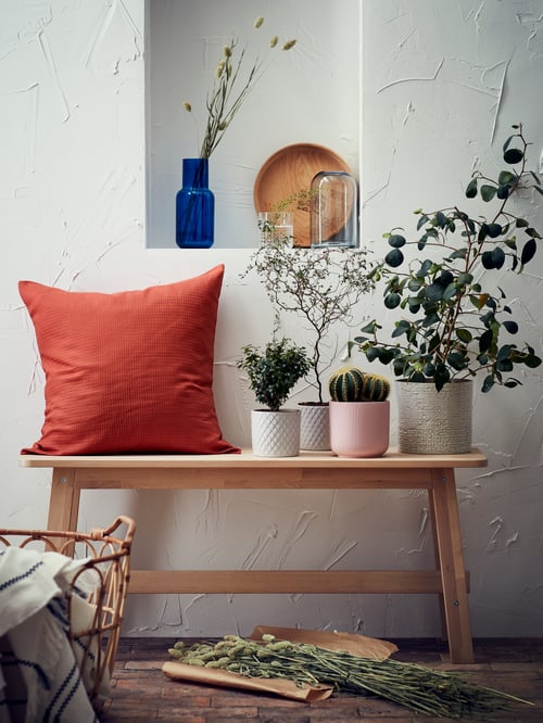 Plusieurs cache-pots avec des plantes et une housse de coussin carrée sur un banc beige. Un vase gris et un vase bleu sont au-dessus.