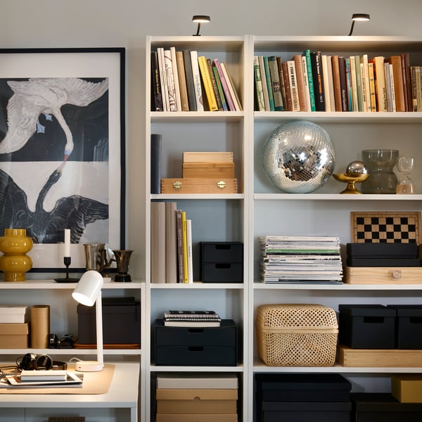 A living room wall with a combination of BILLY bookcases, lit by YTBERG cabinet lighting and filled with books and boxes.