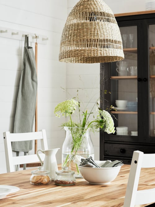 Dining room with a serving bowl, vase, and jar with lid on a wooden table with a pendant lamp shade hanging above.