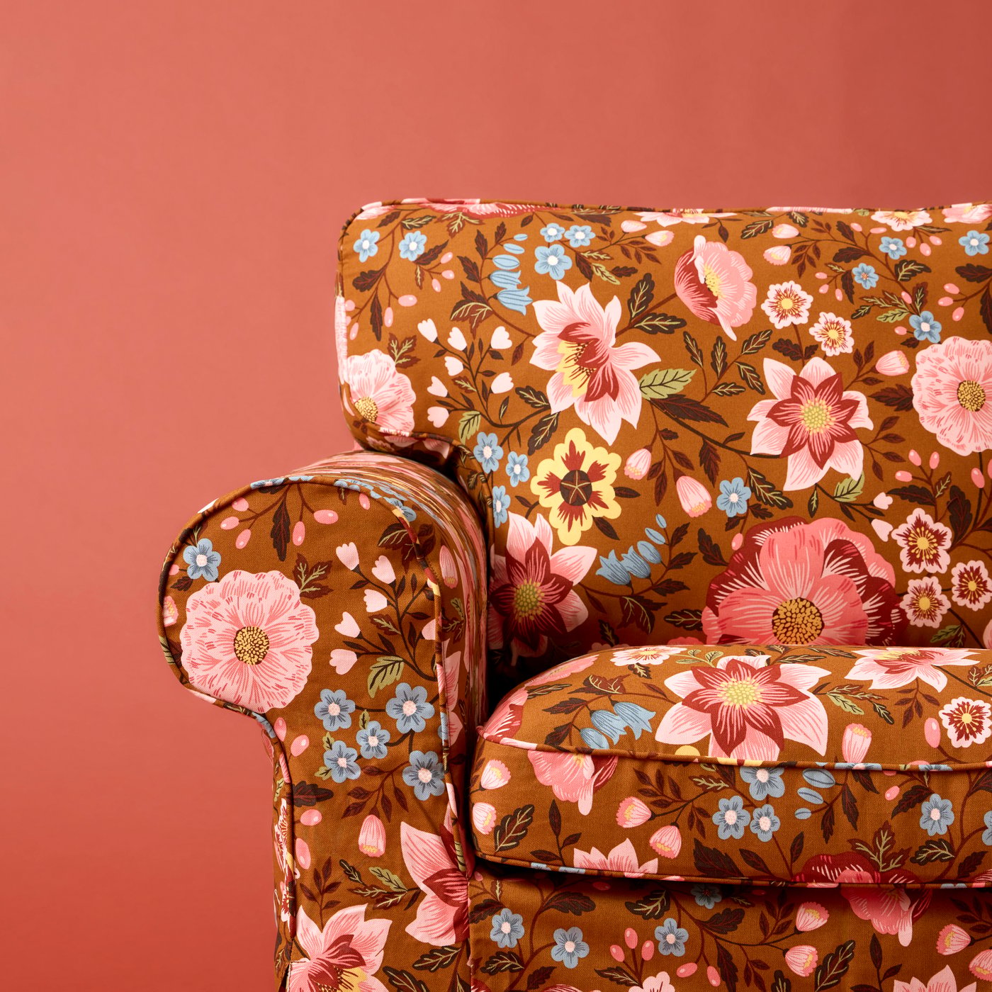 Part of an EKTORP three-seat sofa in a floral-pattern, Mångbyn brown-and-multicolour fabric against an orange-red wall.