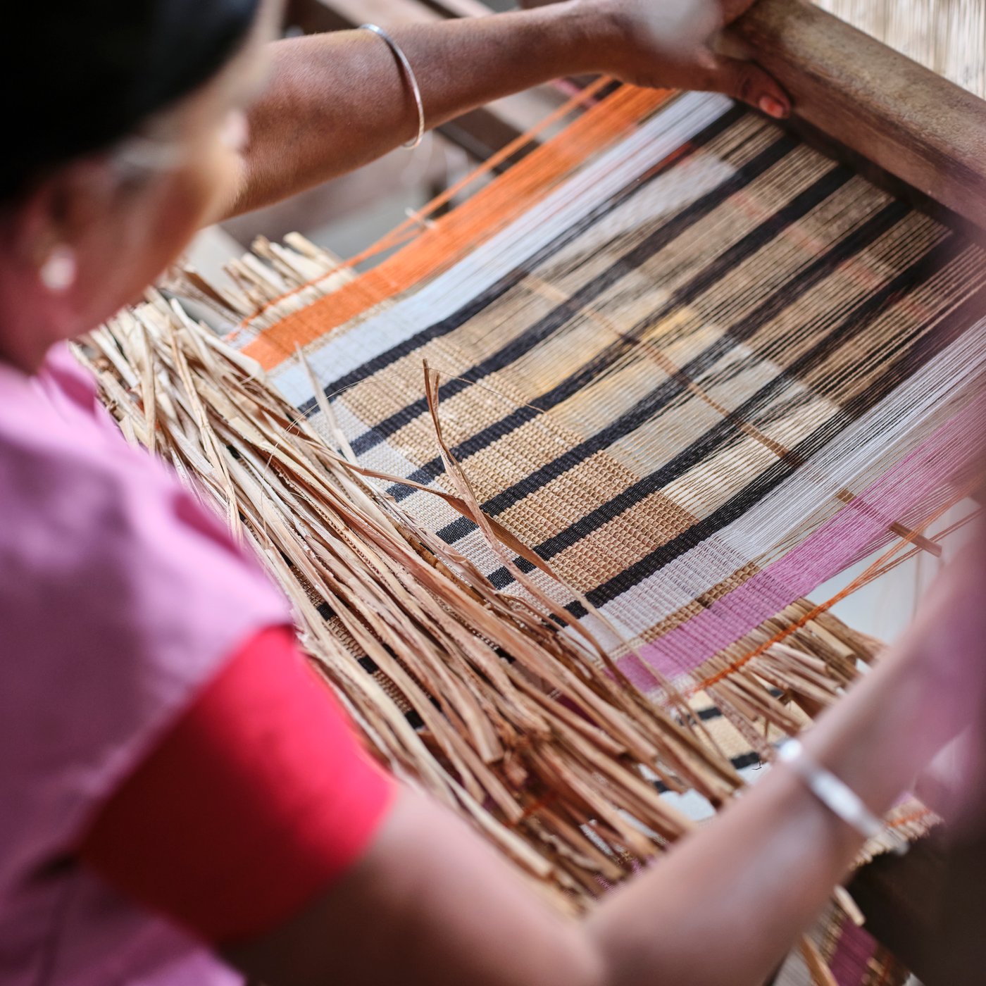 O persoană țese fibre de bananier și fire de bumbac portocalii, albe, negre și roz într-un război de țesut rustic.