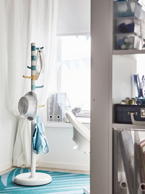 A white clothes stand by a corner with hooks in blue, yellow and green. A grey hat and a blue backpack hang on it.