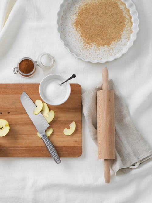Kitchen with tablecloth, chopping board, pie dish, knife, rolling pin, bowl, kitchen towel, apples, spices, teaspoon.