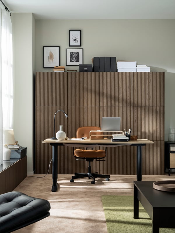 A softly sunlit, carpeted room with a tidy workspace, a wall of wood-front cabinets, a TRULSTORP coffee table and wall art.