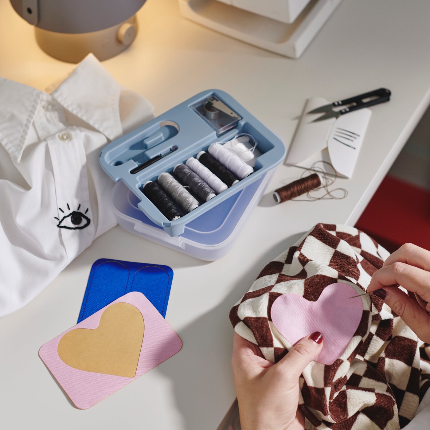 A TRIXIG 39-piece mending set is open on a white desktop near two hands stitching a pink, heart-shaped patch onto a shirt.