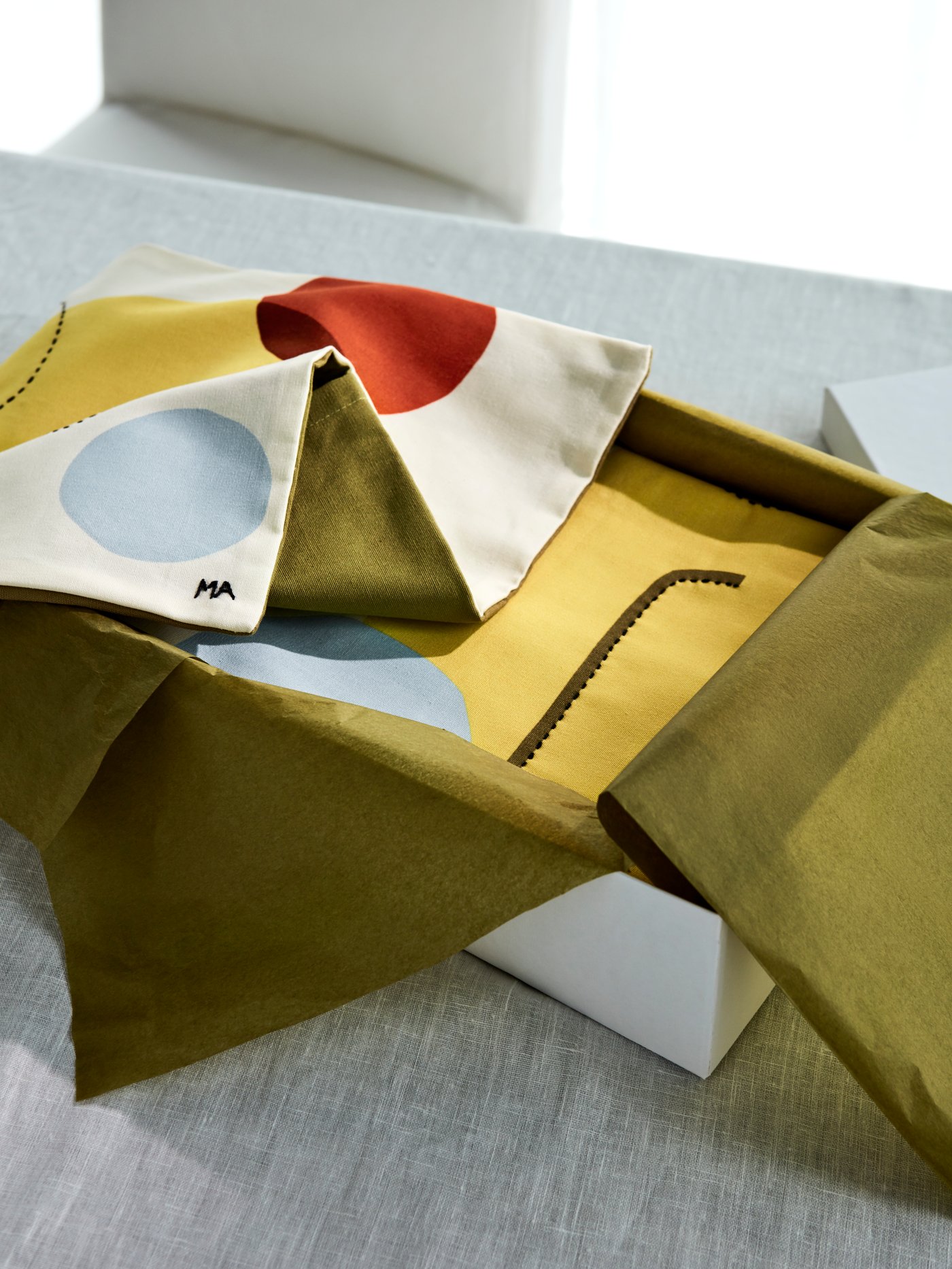 ÄNGSMÄTARE and MANDELPIL cushion covers in abstract patterns in yellow, red, light blue and green wrapped in a gift box.