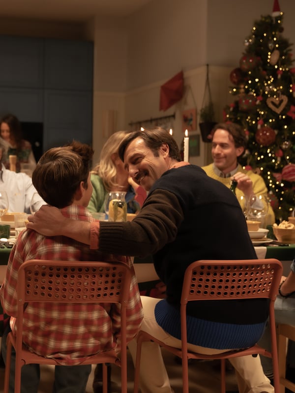 Un uomo seduto a una tavola imbandita per le feste abbraccia con affetto un bambino, sorridendogli. Entrambi sono seduti su sedie rosse modello SANDSBERG.