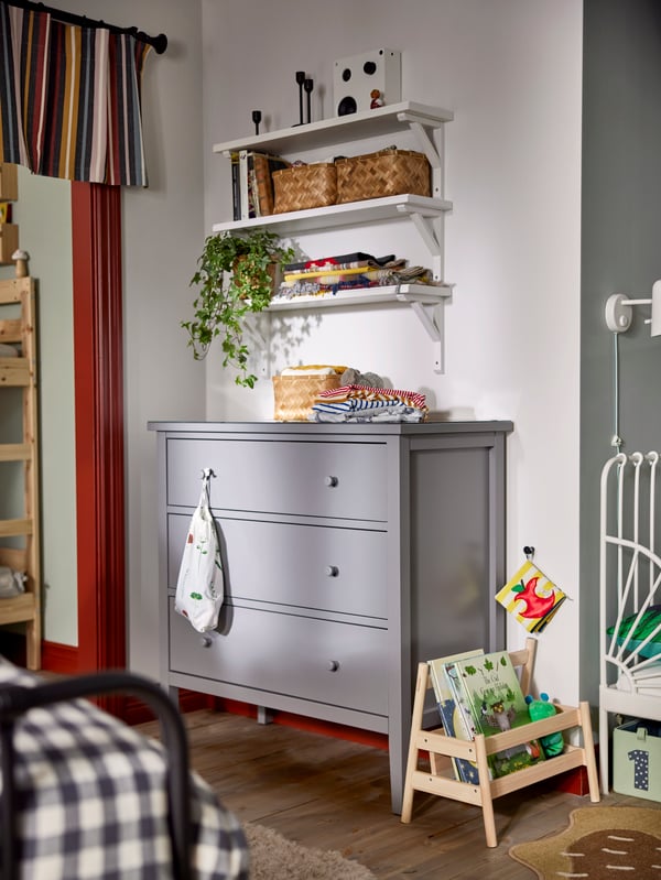 Grey chest of three drawers with three wall mounted shelfs above, baskets, plants and decoration around.