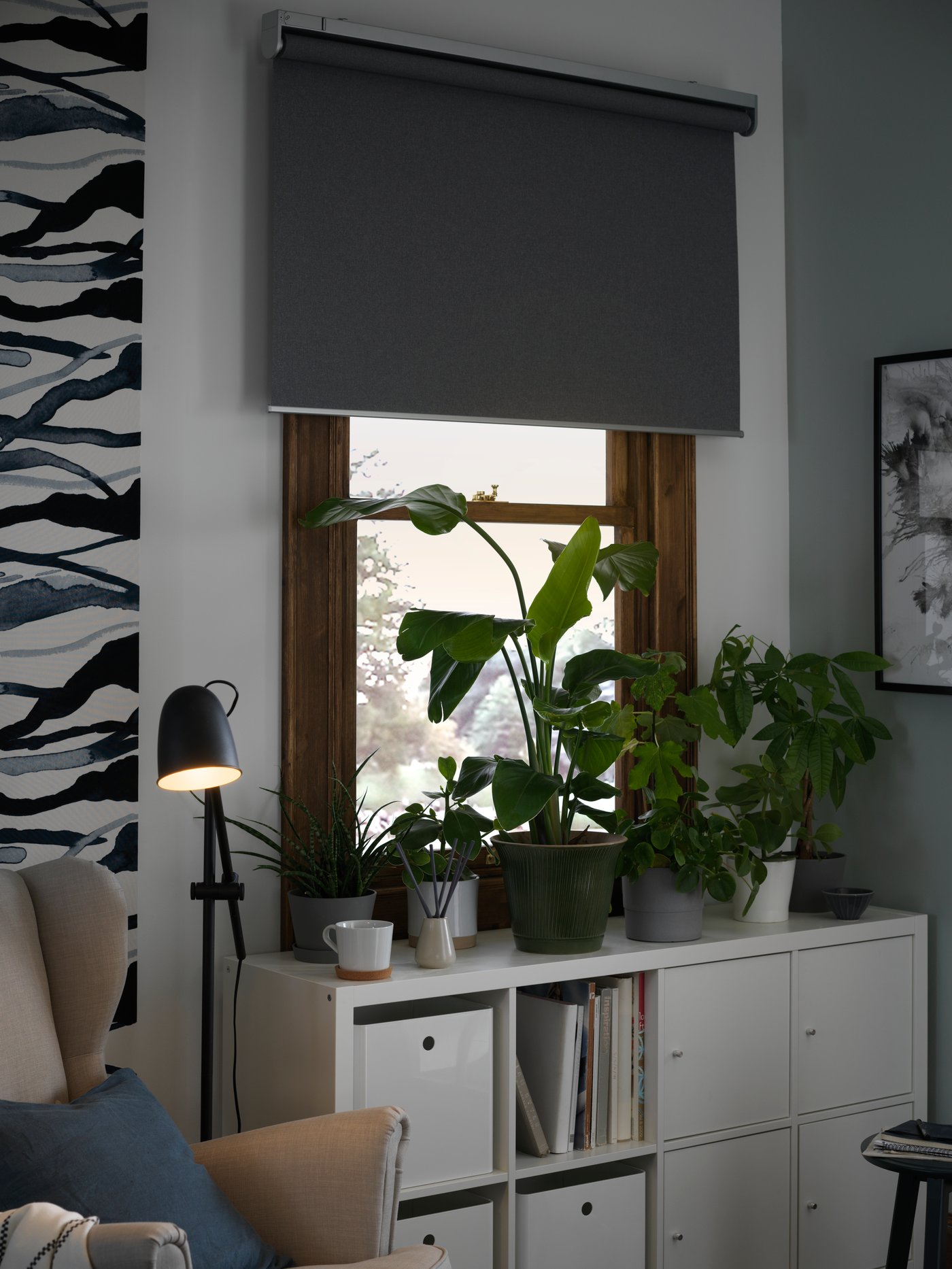 Grey FRYTUR block out roller blind at a window, partly rolled down, a number of potted plants in front.