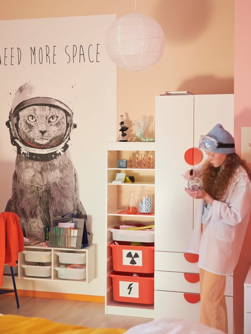 実験用ビーカーを持っている少女が、パステルカラーの部屋の隅にあるホワイトのSMÅSTAD/スモースタード ワードローブ 引き出し付きのそばに立っている。