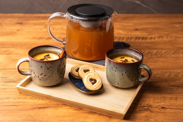 Eine Kanne voller Gewürztee mit zwei Tassen und Keksen auf einem Tisch präsentiert.