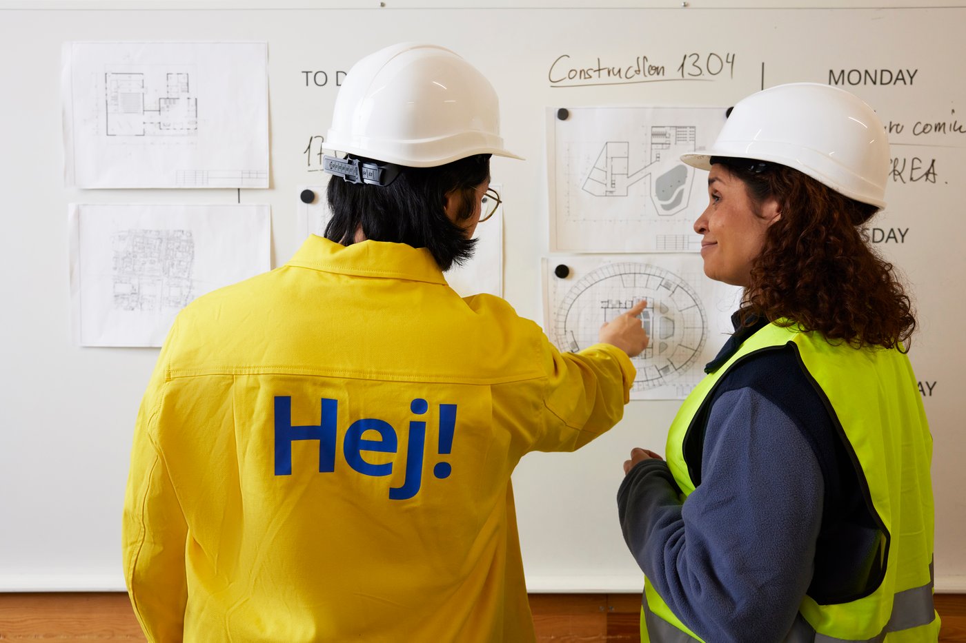 IKEA-työntekijä keltaisessa Hej!-paidassa osoittaa seinällä olevaa suunnitelmakuvaa. Vieressä nainen rakennustyömaakypärä päässä.