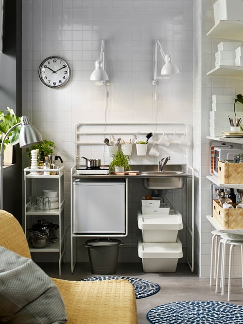 A white SUNNERSTA mini-kitchen has a portable induction hob and a small fridge. A white trolley stores kitchen supplies.