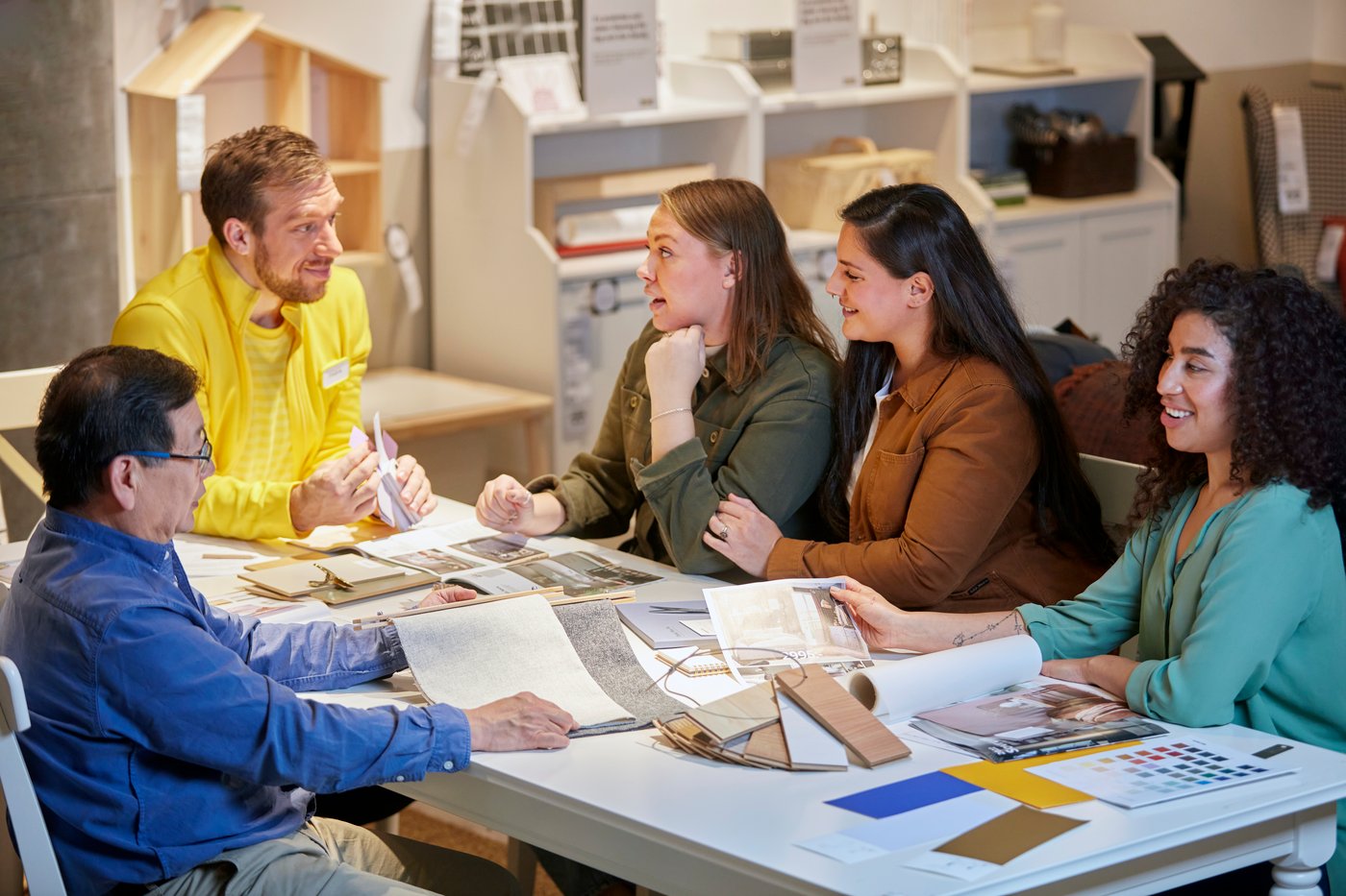 En række mennesker sidder omkring et bord i et IKEA varehus de har åbne kataloger og farveprøver liggende på bordet og en IKEA medarbejder sidder og fortæller.
