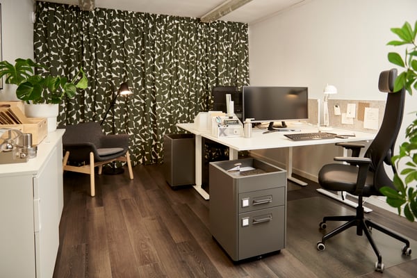 Efterbild på ett modernt kontor med skrivbord, datorskärmar, kontorsstol och växter. Trägolv och mönstrad gardin i bakgrunden.