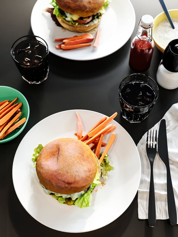 Un plateau de table anthracite avec des assiettes de burgers, des verres de Coca-cola, des serviettes de table blanches, des condiments et des couverts.