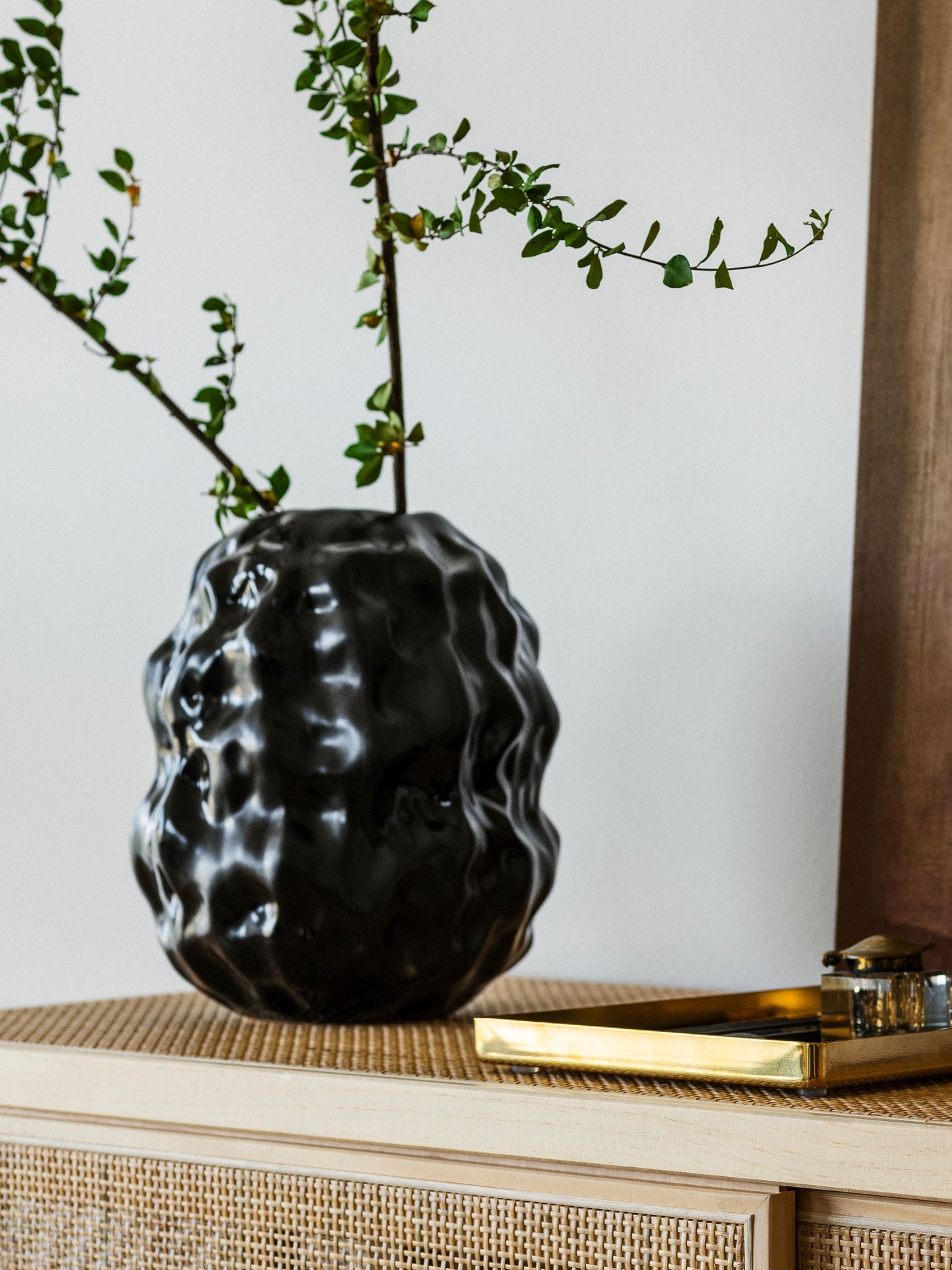 A black STOCKHOLM 2025 vase with a plant sits next to a brass-coloured tray on a pine and rattan cabinet with sliding doors.