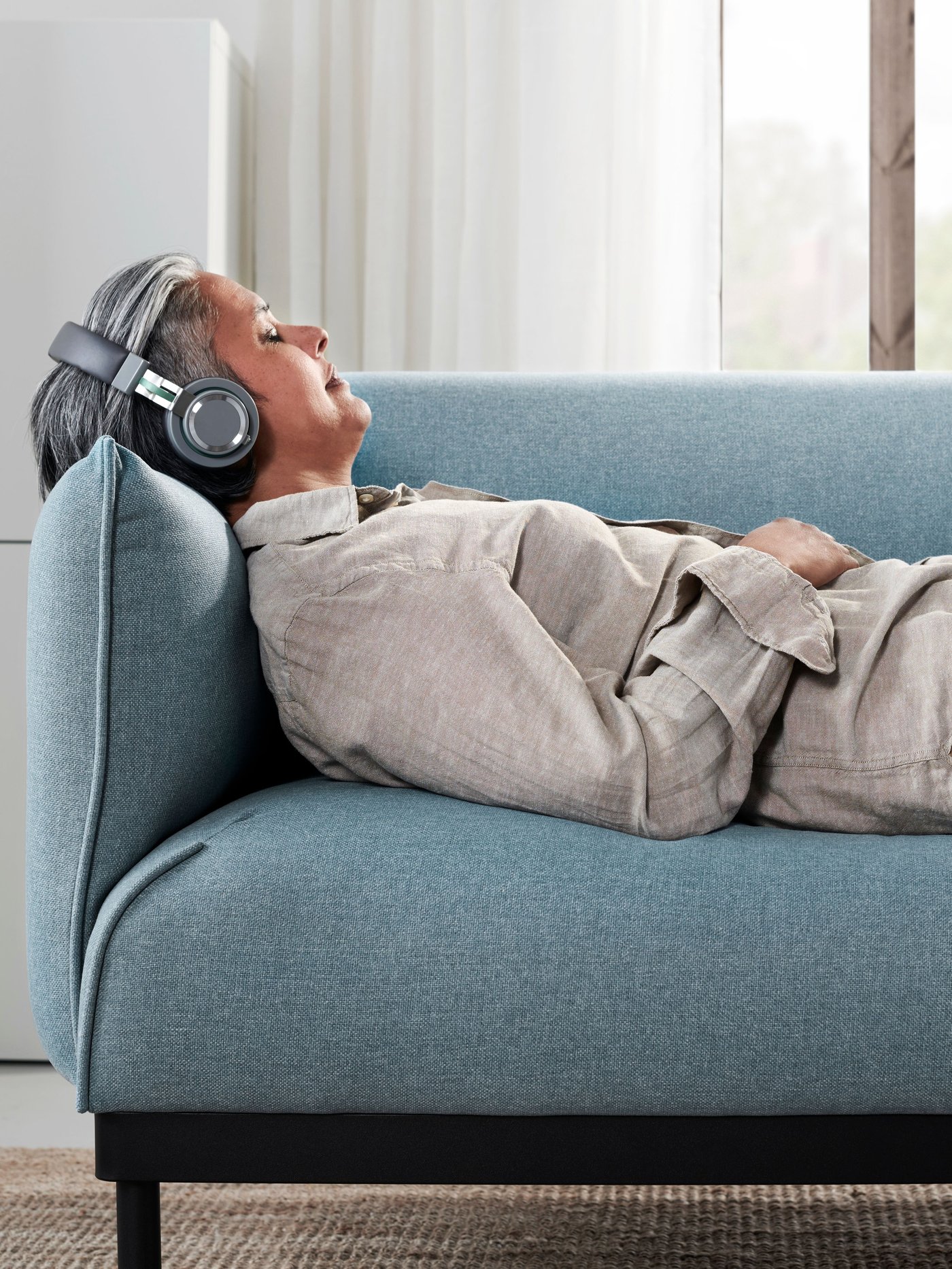 Kobieta z białymi słuchawkami na uszach odpoczywa na jasnoszarej sofie ÄPPLARYD z głową i ramieniem na podłokietniku.