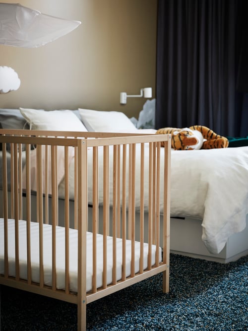 A SNIGLAR cot next to a double bed which has a DJUNGELSKOG soft toy on it. There is a VINDUM high pile rug on the floor.