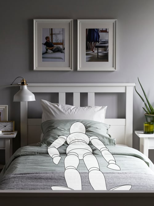 A white HEMNES bed with a cartoon drawing of a person sleeping on it. Above the bed there is an off-white RANARP work lamp.