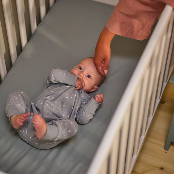 A baby lies in a SUNDVIK cot that has a blue GULDVÄVARE sheet on the mattress, with an adult’s hand stroking its head.