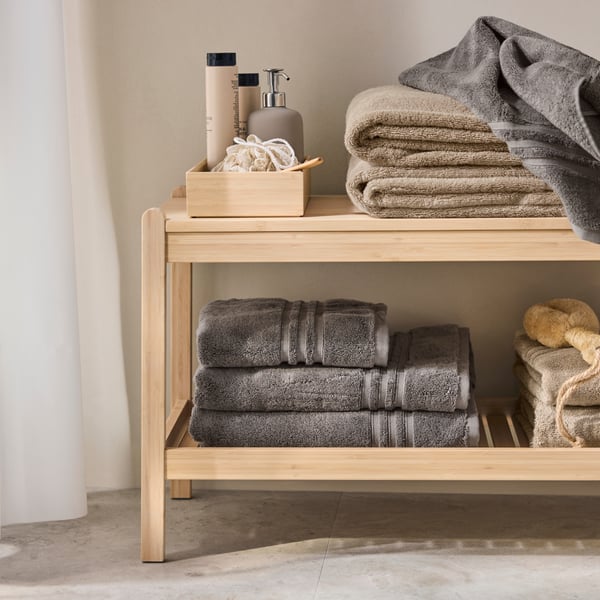 A bathroom with a light wooden shelf displaying neatly stacked grey BROKGLIM bath towels, along with toiletries.