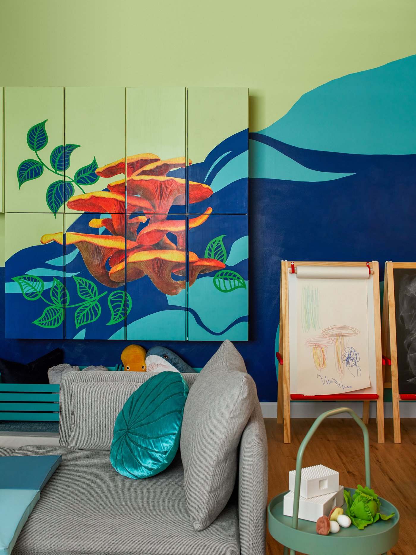 A storage is on the wall and there is painting of chanterelles on a green and blue background. There is a grey sofa in front of them with a green coffee table full of toys. 