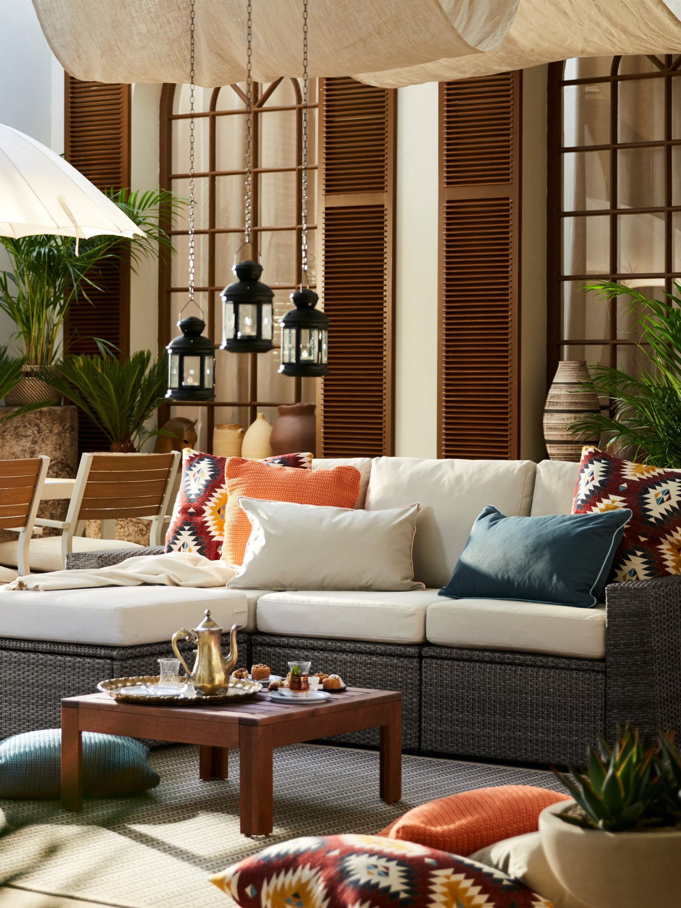 En udendørs SOLLERÖN udendørs modulopbygget sofa med en hvid fodskammel og forskellige puder slænget ovenpå står på en tarrasse under en parasol.