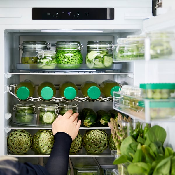 A person is reaching for a jar of pickles from a shelf in an integrated SKOGNÄS fridge/freezer, full of stored food items.