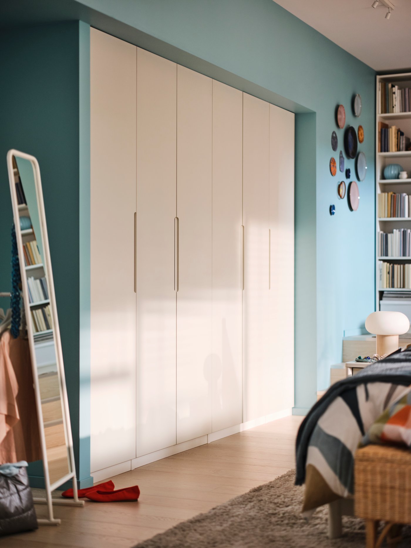 A line of wardrobes in a niche along a blue wall, with a mirror to the left and the corner of a bed in the foreground.
