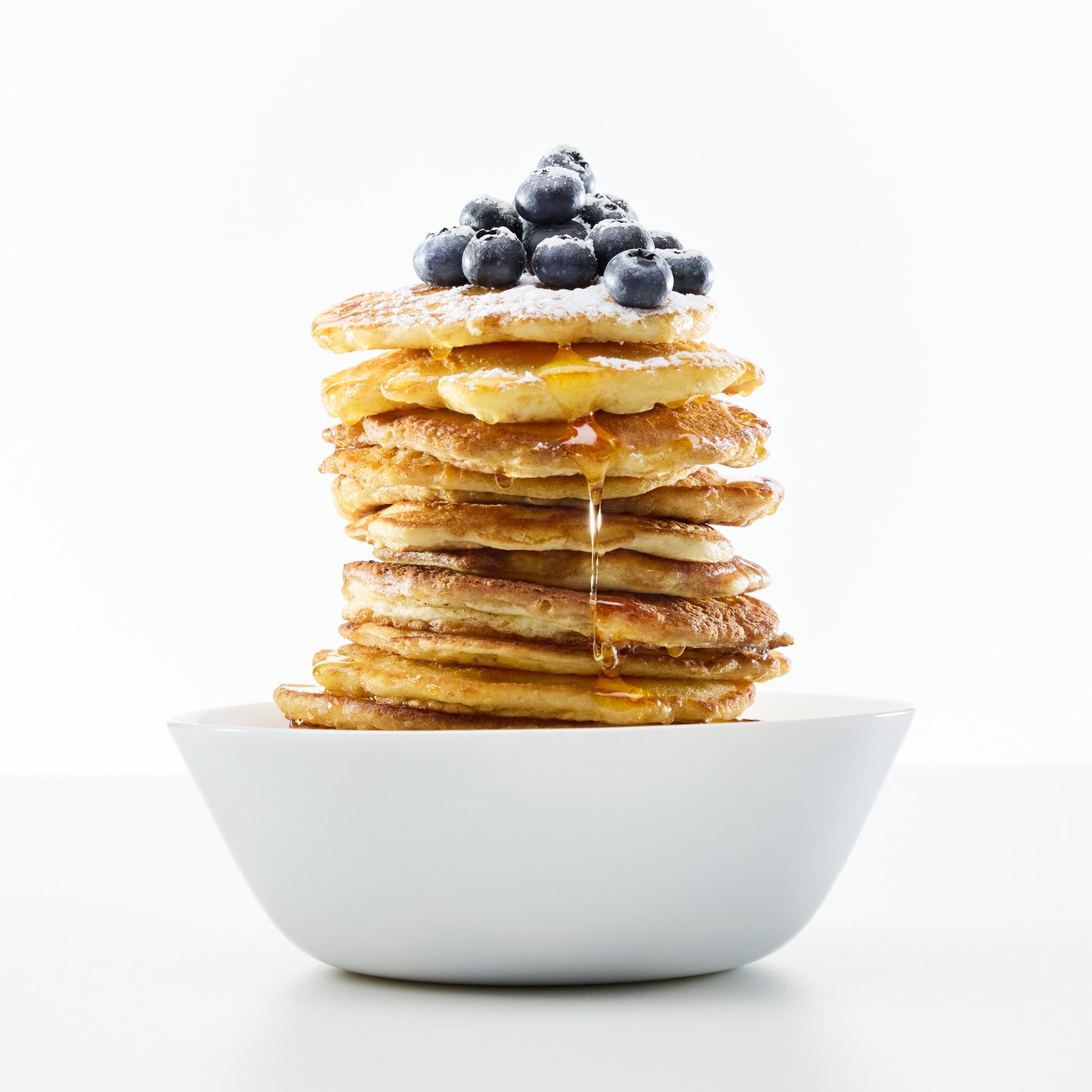 A white OFTAST bowl, filled with a tall stack of maple syrup-drenched pancakes with blueberries on top.