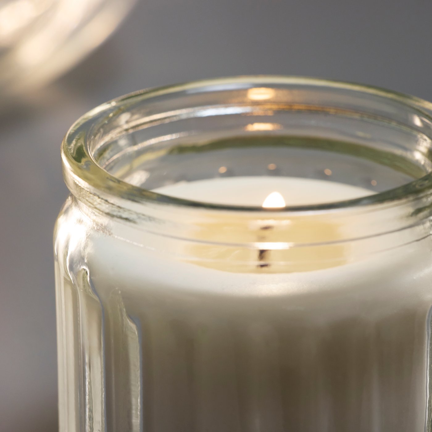 Primo piano di una candela profumata color beige chiaro accesa, in un vasetto di vetro trasparente.