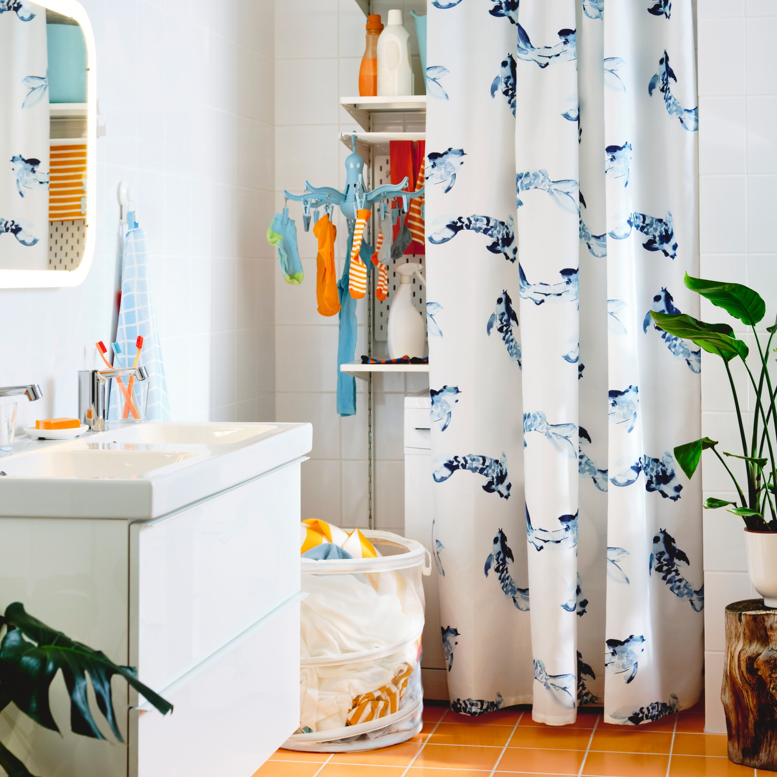 Un baño con muros blancos y piso de baldosas naranjas, con un vanitorio blanco GODMORGON. Al fondo y detrás de una cortina blanca con diseños azules se ve un espacio de lavado con ropa colgada y un canasto con ropa sucia.