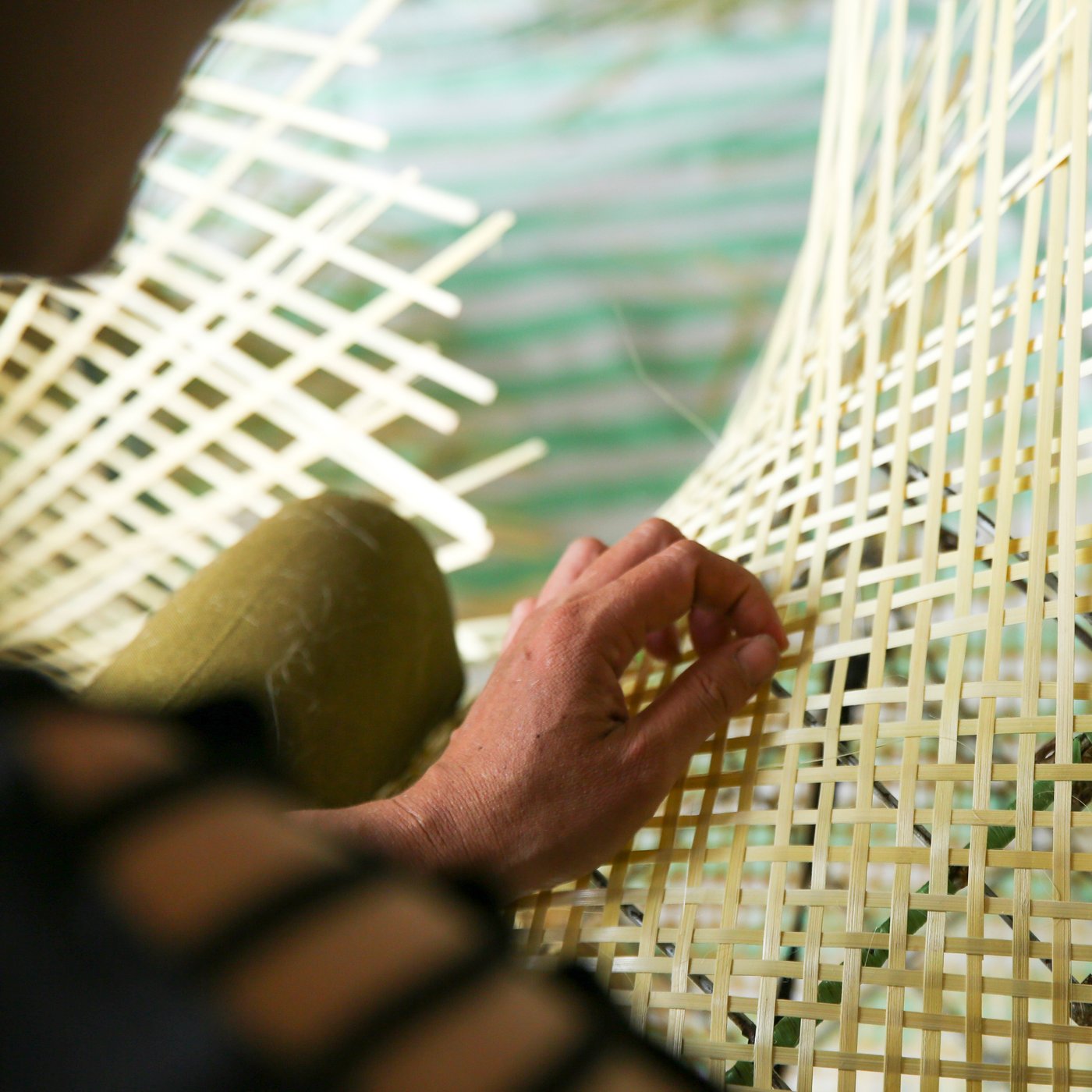 Un artisan avec un pantalon vert qui tresse à la main un maillage de bambou jaunâtre aéré sur un pied de lampe métallique.