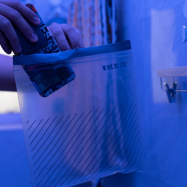 A man in a bathroom puts a cellphone into an ISTAD resealable plastic bag. The lighting is blue.