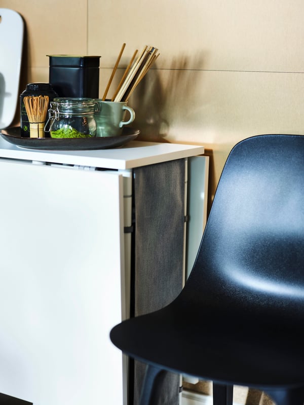 A folded up white/light grey KALLHÄLL gateleg table with storage against a wall, next to an anthracite ODGER chair.