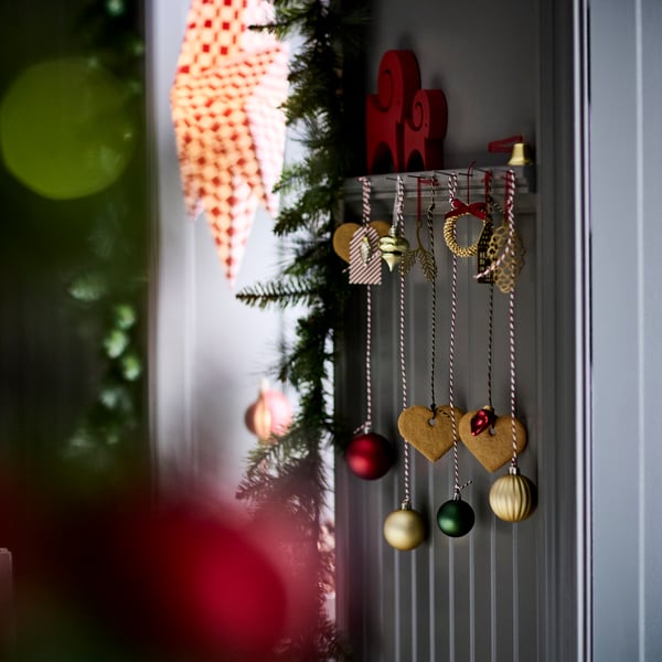 En festlig julscen med dekorerad vägg med hängande prydnader – gyllene hjärtan, röda och gröna kulor samt intrikata gulddetaljer. I bakgrunden syns en julgranskvist och suddiga ljus.