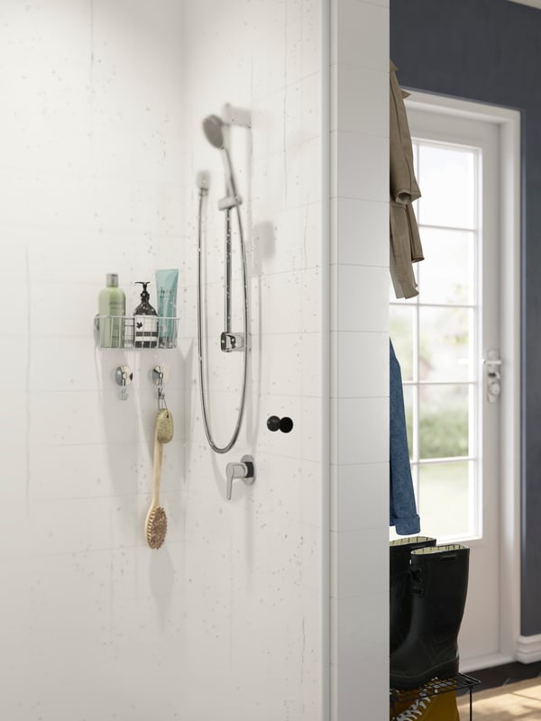 A shower is attached to a wall in a corner of a mudroom. There is a shelf with bathroom products and a brush on the wall.