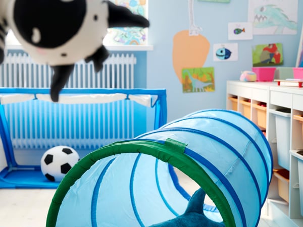 Blue DVÄRGMÅS play tunnel on the floor of a children’s room, with BLÅHAJ soft toy inside it and a hand puppet in foreground.