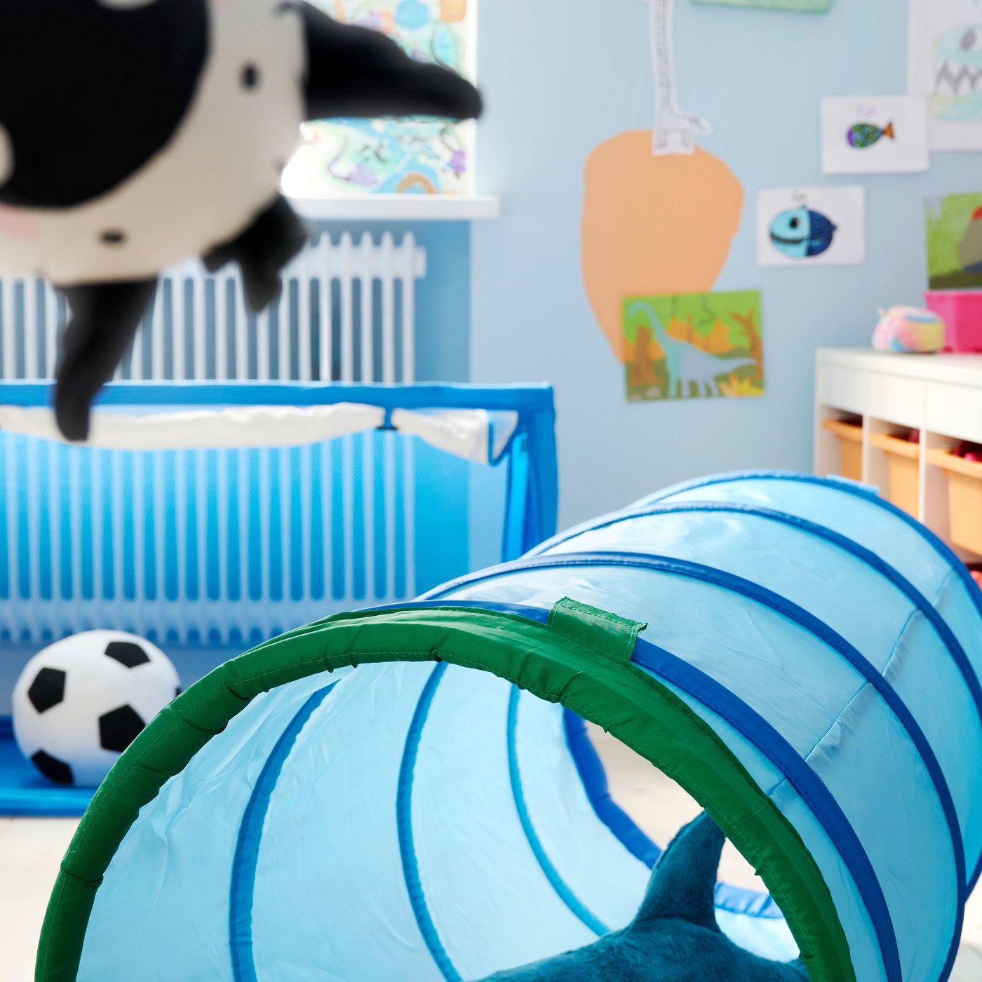 Blå DVÄRGMÅS lektunnel på golvet i ett lekrum för barn, med mjukdjuret BLÅHAJ inuti och en handdocka i förgrunden.
