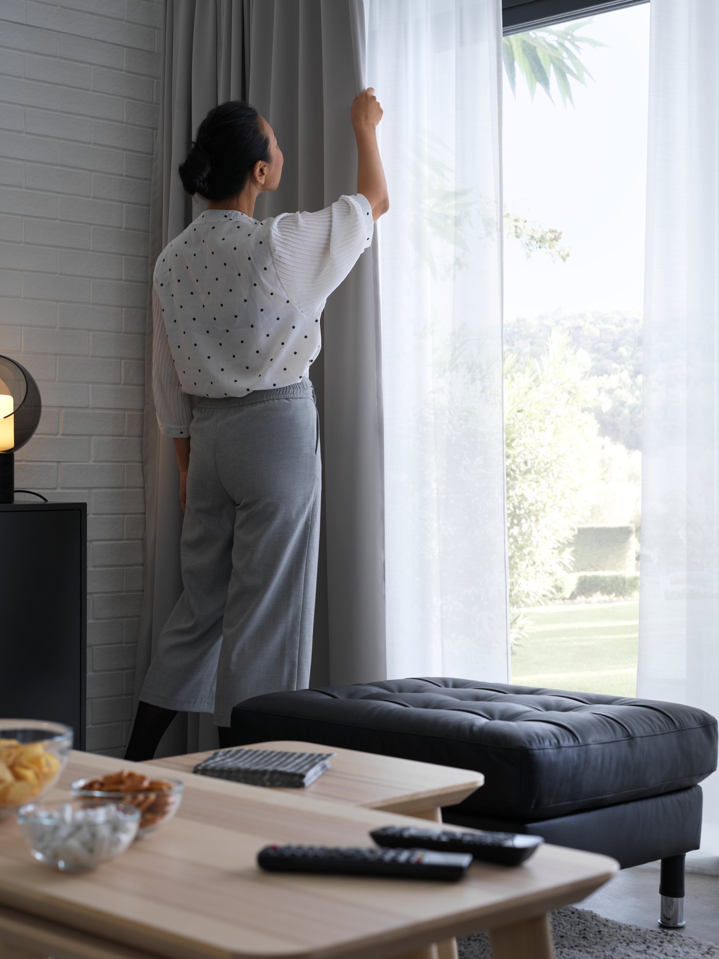 Une femme ajuste des rideaux dans un salon, avec une ottomane LANDSKRONA noire et des tables LISABO.